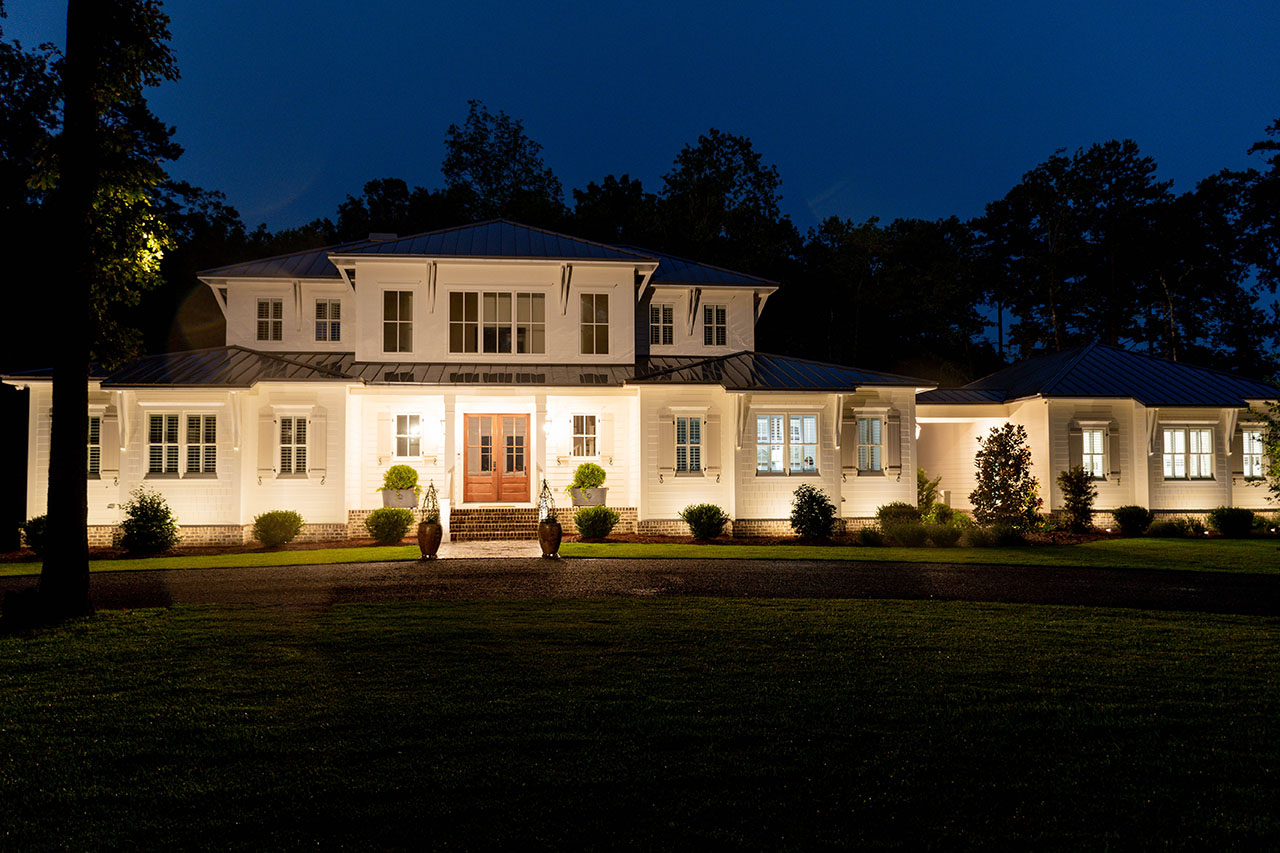 A large, well-lit two-story house with white exterior walls, multiple windows, and a manicured lawn is shown at night. Two figures are standing near the front entrance illuminated by lights.